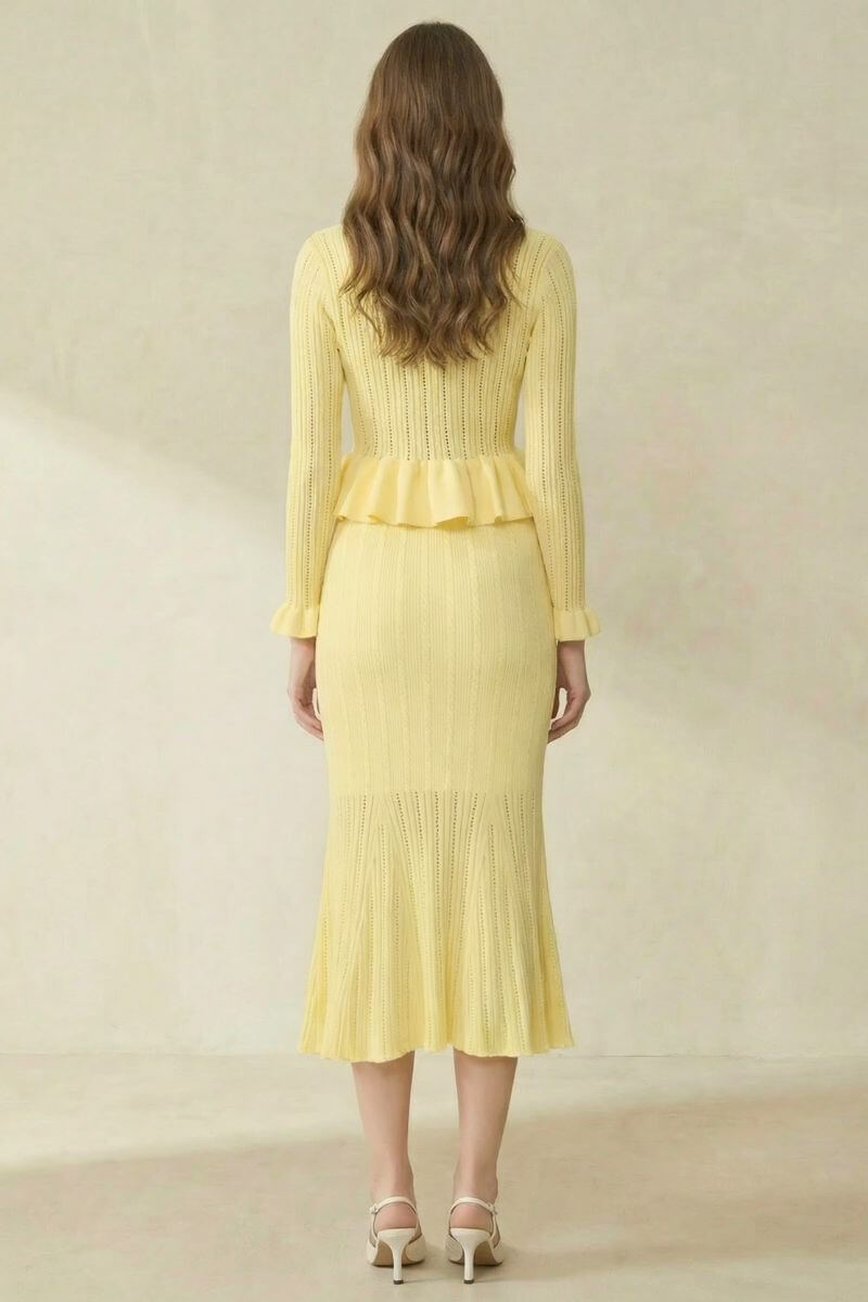 A woman wearing a soft yellow knit two-piece set with a peplum cardigan and flared midi skirt, shown from the back in a minimal studio setting.