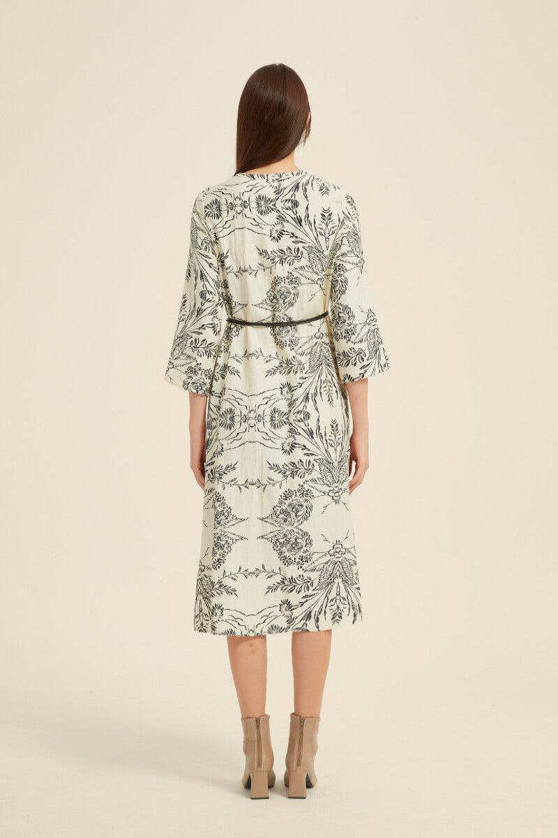 Back view of a cream midi dress featuring an intricate black botanical print, wide sleeves, and a thin black waist belt.