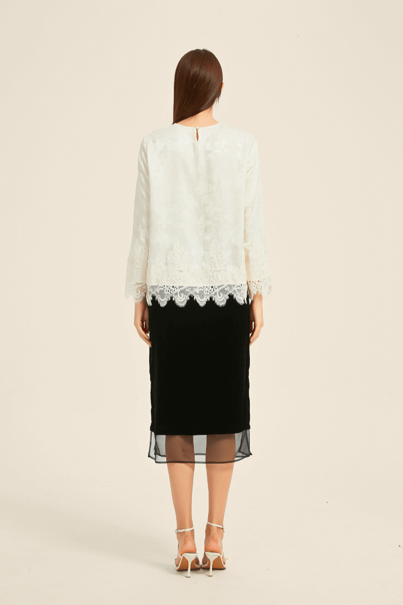 Back view of a woman wearing an ivory lace-trim long-sleeve blouse and a black midi skirt with sheer hem, styled with white strappy heels.