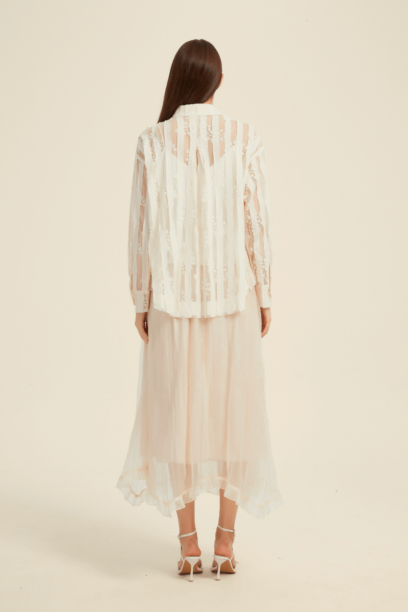 Back view of a model wearing an ivory sheer stripe blouse with a collared neckline and long sleeves, paired with a flowy ivory midi skirt featuring a ruffled hem.
