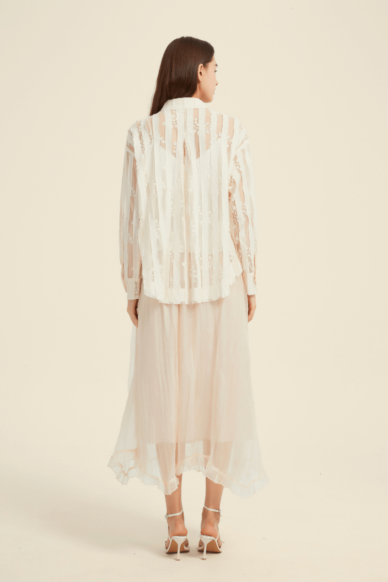 Rear view of a woman wearing a cream sheer lace blouse with vertical embroidery, paired with a flowy, asymmetrical blush midi skirt.
