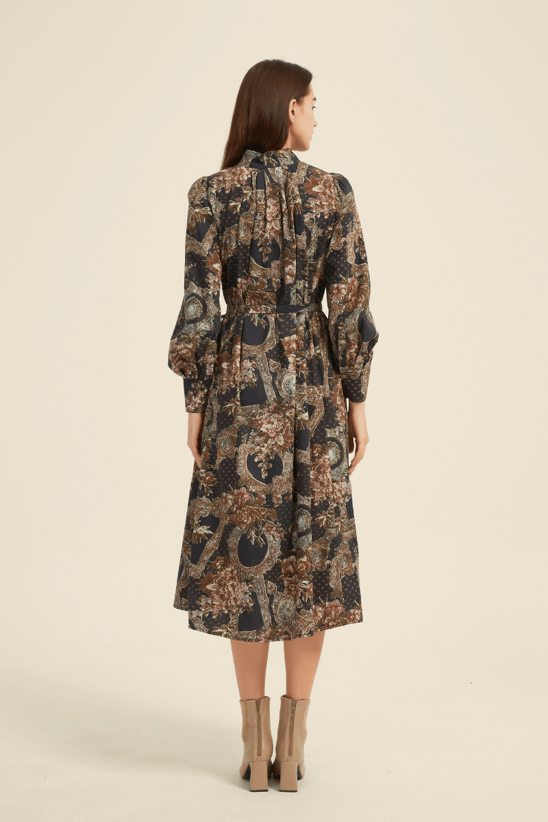 Back view of a vintage-inspired midi dress featuring an intricate floral and geometric print in earthy tones, long puff sleeves, a high neckline, and a belted waist for a flattering silhouette.