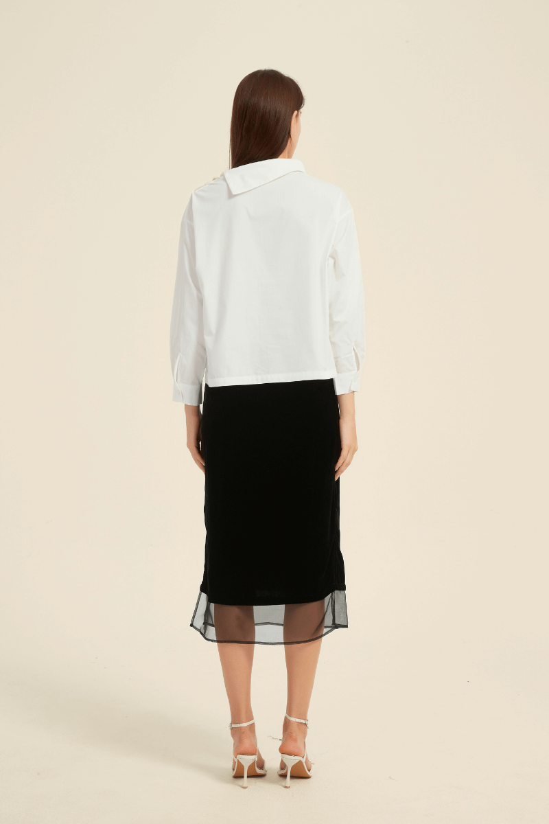 Back view of a woman wearing a white long-sleeve blouse with an asymmetric collar, paired with a black midi skirt with sheer hem panels and side slits.