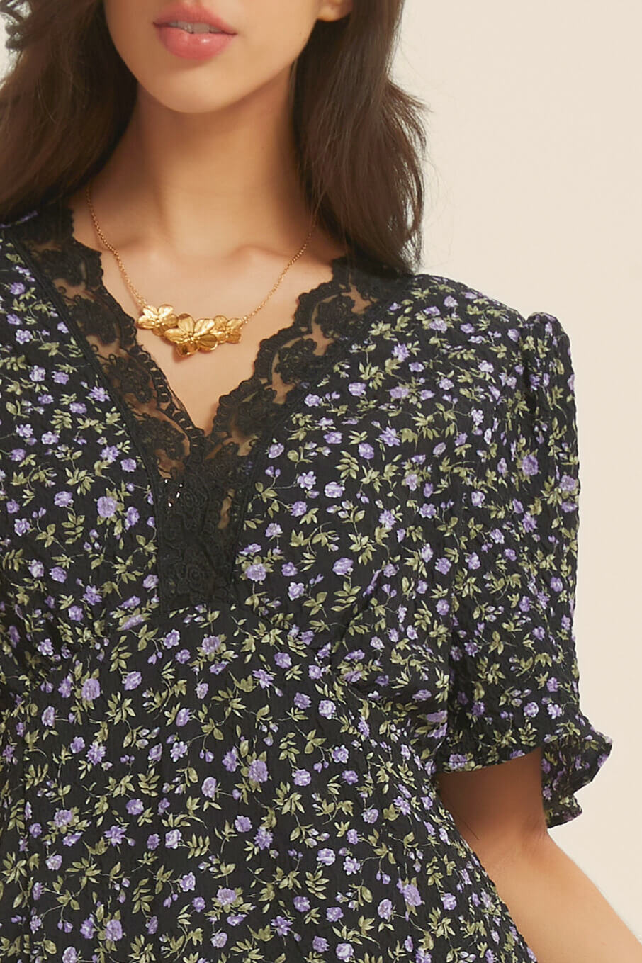 Close-up of black dress with purple floral print and sheer lace-trimmed neckline, showcasing puff sleeves and delicate gold floral necklace.