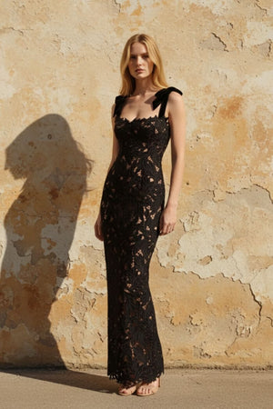 model wearing a black lace bow-strap evening gown in natural sunlight against a textured stone wall, showcasing a refined fitted silhouette.