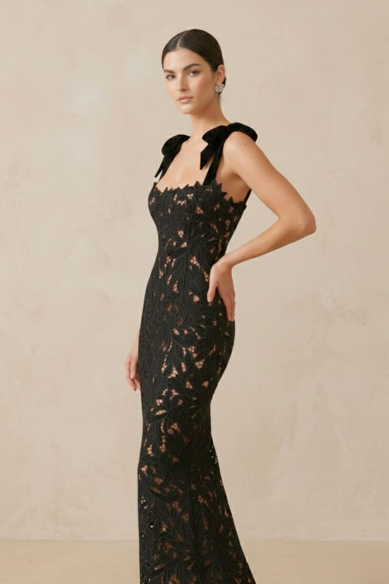 side view of a fitted black lace evening gown with velvet bow straps and scalloped neckline, model posing in a beige background.