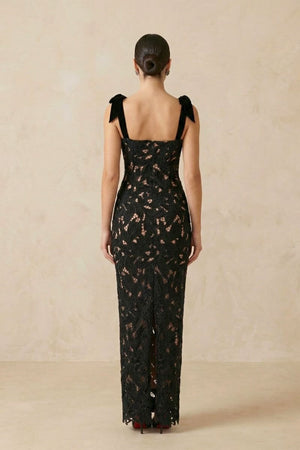 back view of a black lace evening gown with velvet bow straps, fitted shape, and back slit, photographed in a beige studio.