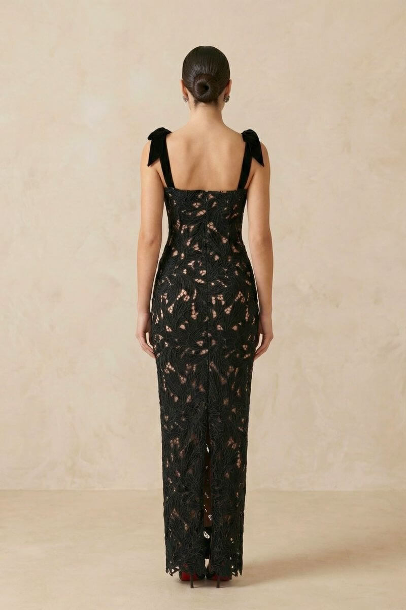 back view of a black lace evening gown with velvet bow straps, fitted shape, and back slit, photographed in a beige studio.