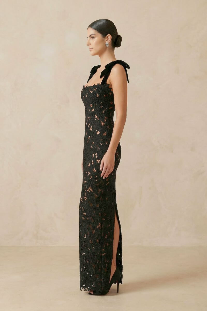 side profile of a model in a black lace gown with velvet bow straps and thigh-high slit, shown in a neutral-toned studio.