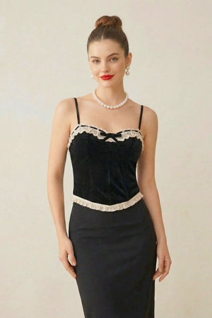 Model wearing a black velvet corset top with cream lace trim and thin straps, styled with pearls and red lipstick in a studio setting.