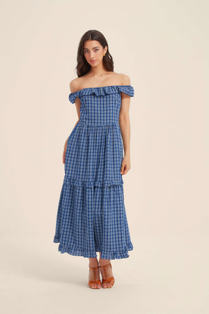 Woman wearing a blue off-shoulder gingham maxi dress with ruffle trim, perfect for summer getaways or outdoor brunches.