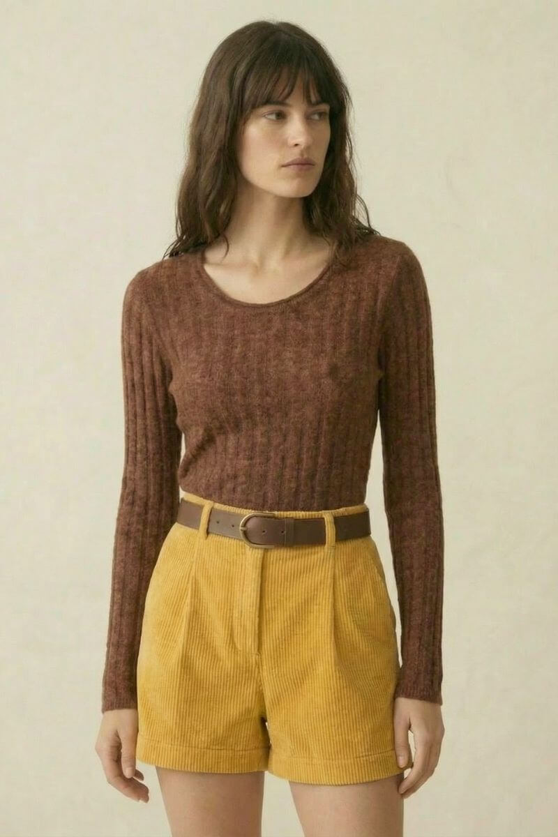 A woman wearing a brown ribbed long-sleeve sweater styled with high-waisted yellow corduroy shorts and a brown belt, standing against a neutral beige backdrop.