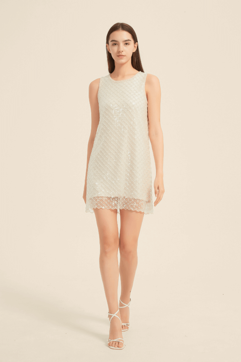 Model walking in a sleeveless champagne mini dress with shimmering sequin embellishment in a diamond pattern, round neckline, and delicate scalloped hem, styled with white strappy heels for an elegant party look.