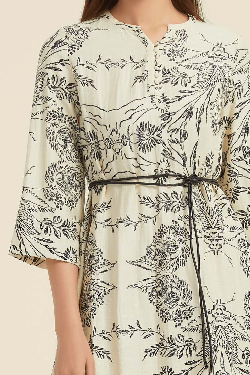 Close-up of a cream midi dress with a black botanical print, cloth-covered buttons at the neckline, and a slim black waist tie.
