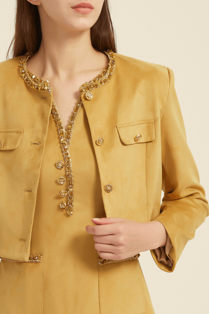 Close-up view of a mustard yellow two-piece set showcasing a sleeveless mini dress with beaded trim neckline and button accents, paired with a cropped long-sleeve jacket featuring flap pockets and beaded trim.