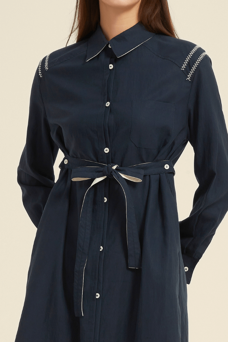 Close-up view of a navy midi shirt dress featuring a collared neckline, button-down front, belted waist, and contrast stitching on the shoulders.