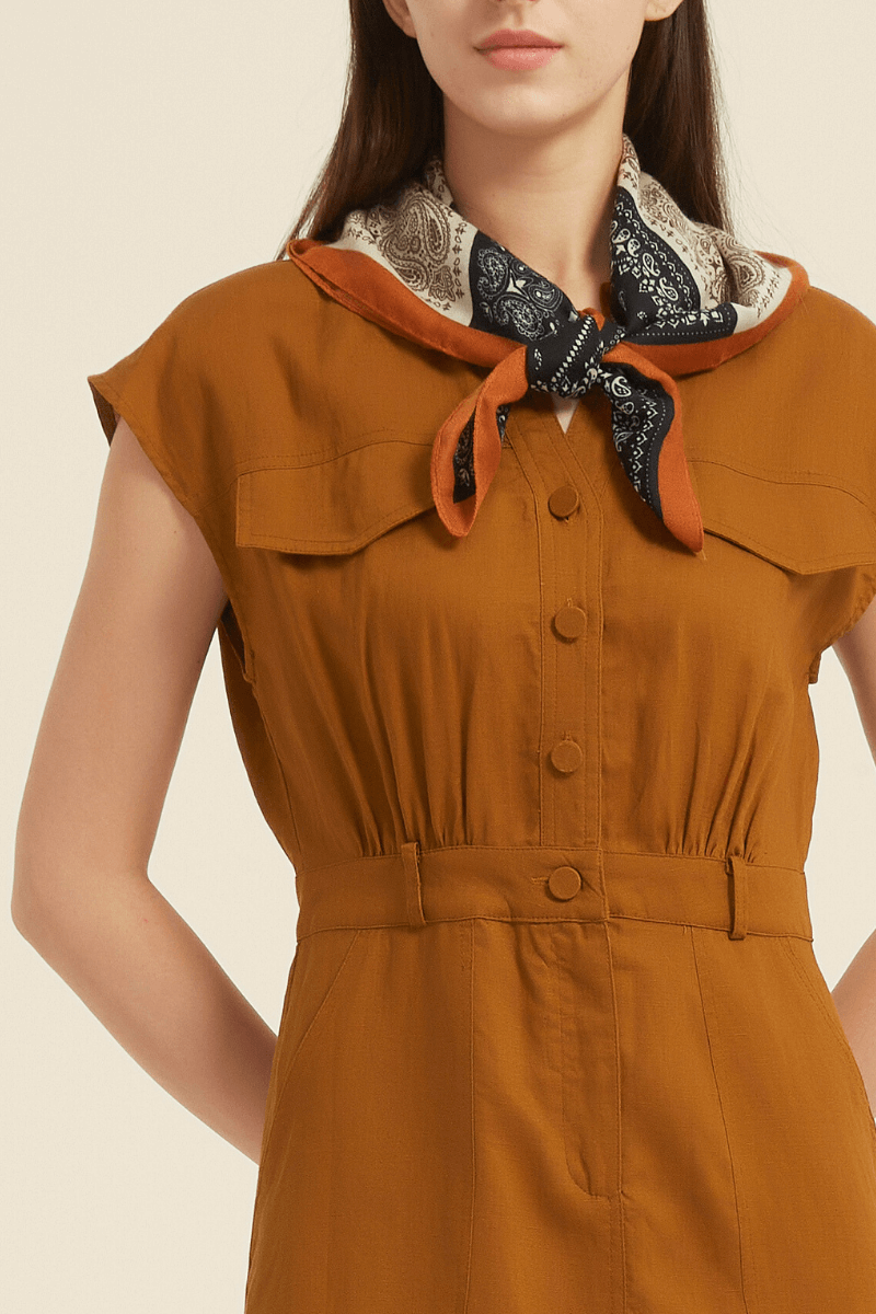 Close-up view of a rust cap sleeve midi shirt dress featuring a button-down front, elastic waist, chest flap pockets, and a printed scarf tied at the neckline.