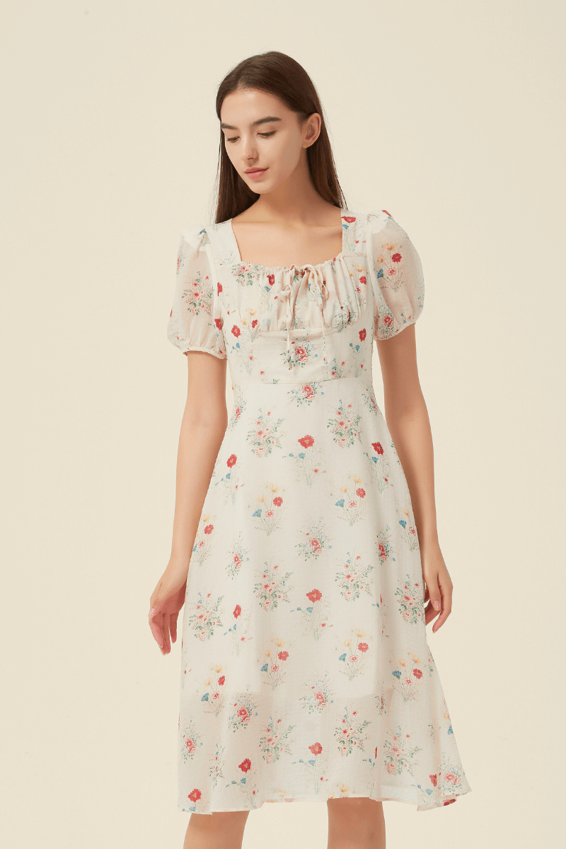 Model wearing a cream midi dress adorned with a scattered floral print in red, yellow, and blue, featuring a square neckline with gathered tie detail, short puff sleeves, and a softly flared skirt for a vintage-inspired silhouette.