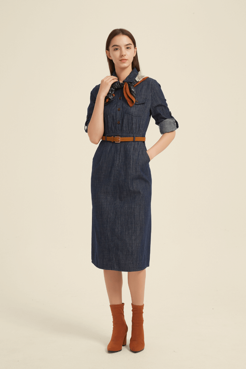 Woman wearing a dark denim button-down midi dress with collared neckline, belted waist, rolled-up sleeves, and a chic tailored fit.