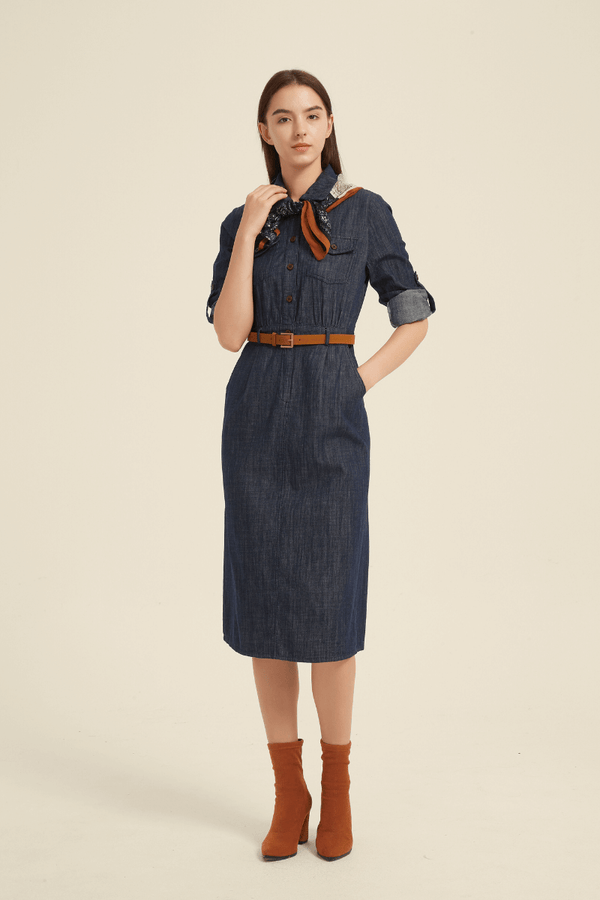 Woman wearing a dark denim button-down midi dress with collared neckline, belted waist, rolled-up sleeves, and a chic tailored fit.