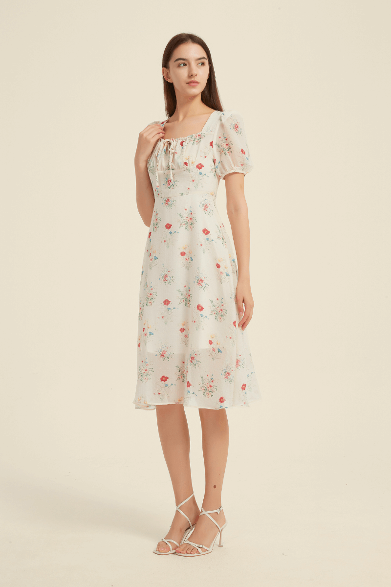 Model wearing a cream midi dress with a delicate red and pink floral print, featuring a square neckline with tie detail, short puff sleeves, and a softly flared hemline for a romantic, vintage-inspired look.