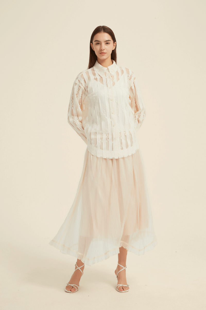 Front view of a model wearing a sheer lace striped blouse with a collared neckline and long sleeves, paired with a flowing ivory midi skirt and strappy sandals.