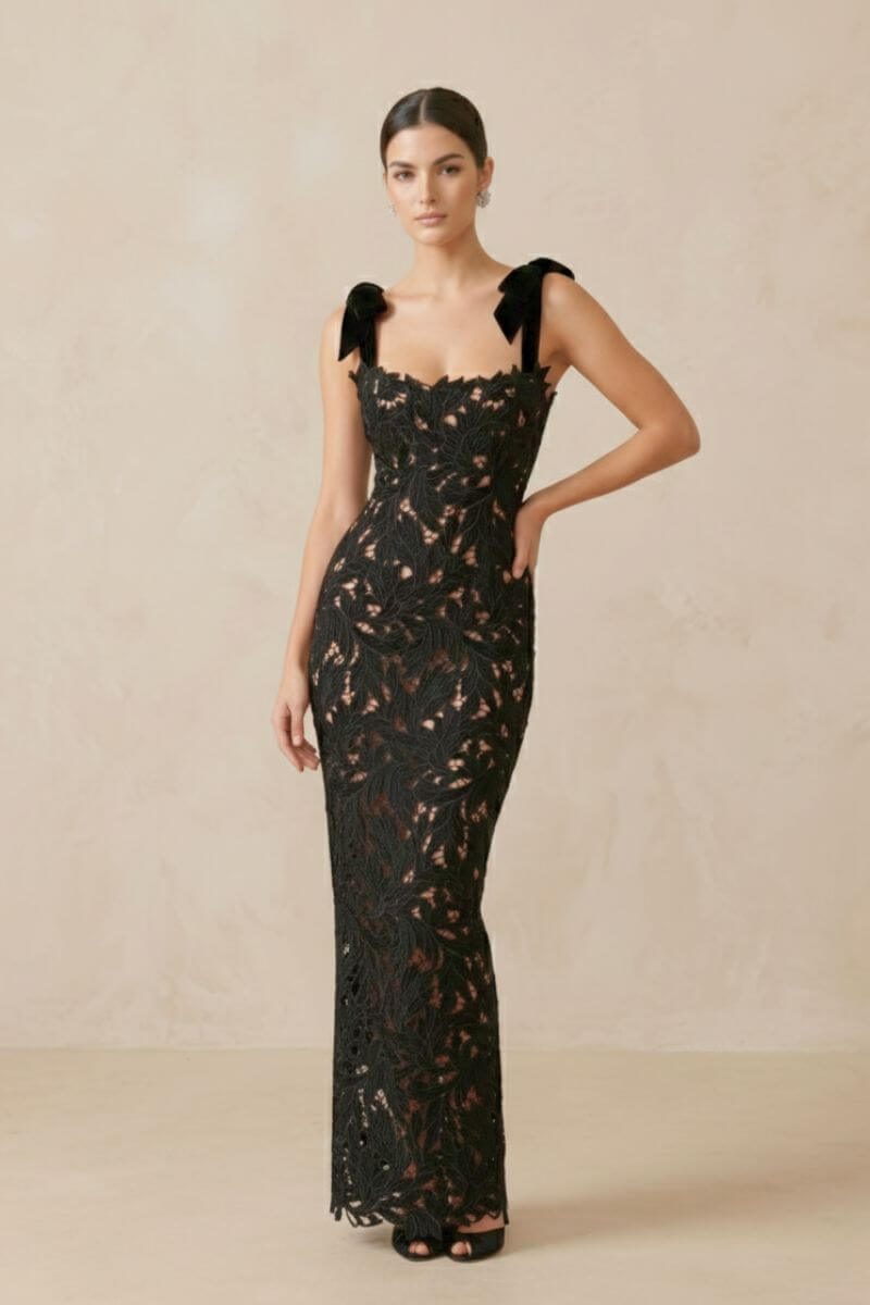 model wearing a full-length black lace gown with velvet bow straps and fitted silhouette, standing in a neutral beige studio.