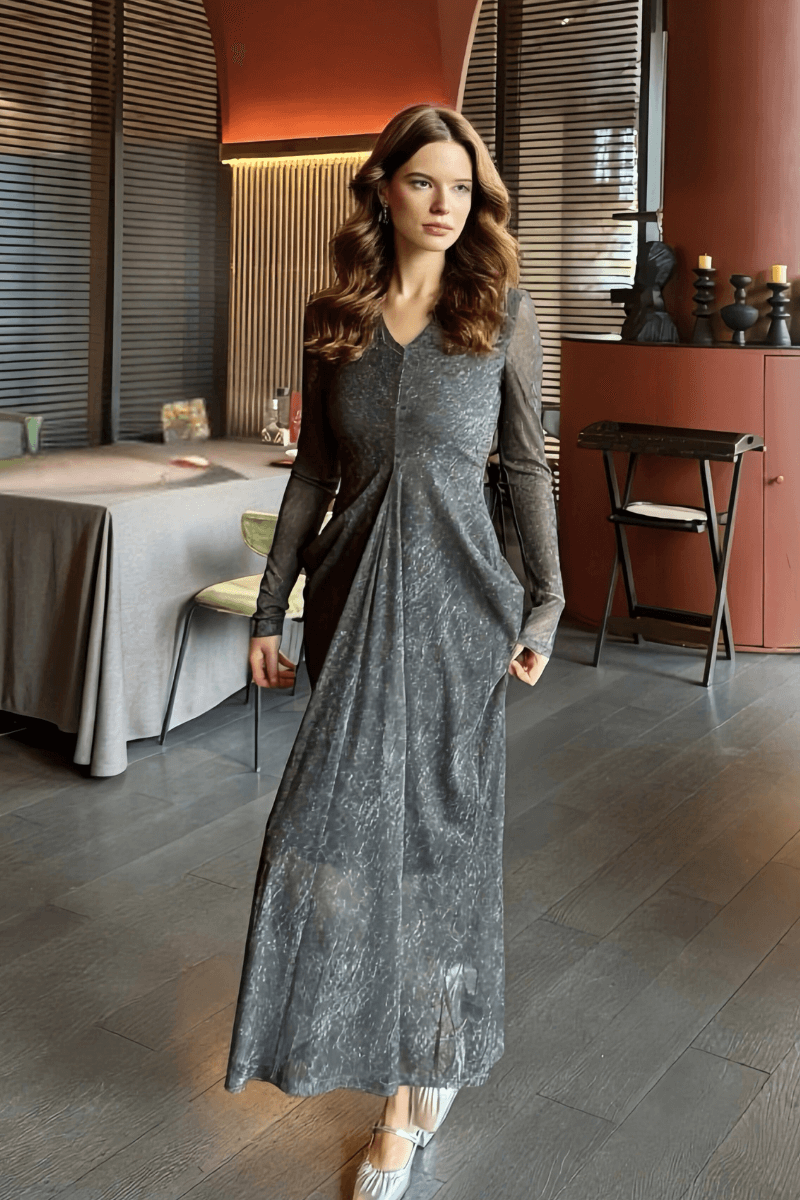 Woman wearing a grey maxi dress with long sheer sleeves, subtle textured pattern, and a v-neckline, styled with silver heels in a modern indoor setting.