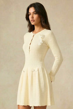 Side profile of an ivory knit mini dress featuring front button detailing and shoulder floral appliqués against a neutral studio backdrop.
