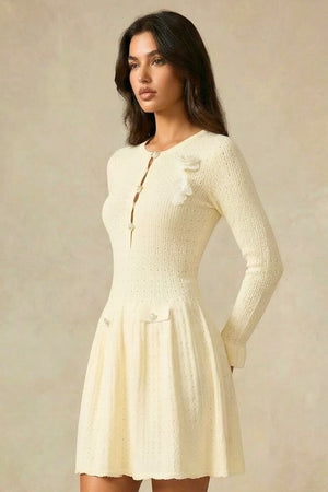 Side profile of an ivory knit mini dress featuring front button detailing and shoulder floral appliqués against a neutral studio backdrop.