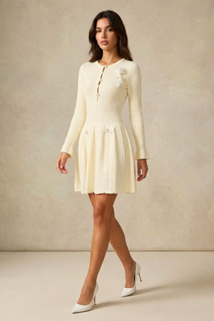 Full-body walking shot of a woman in an ivory knit mini dress with pearl-style buttons and a flared skirt, paired with white heels.