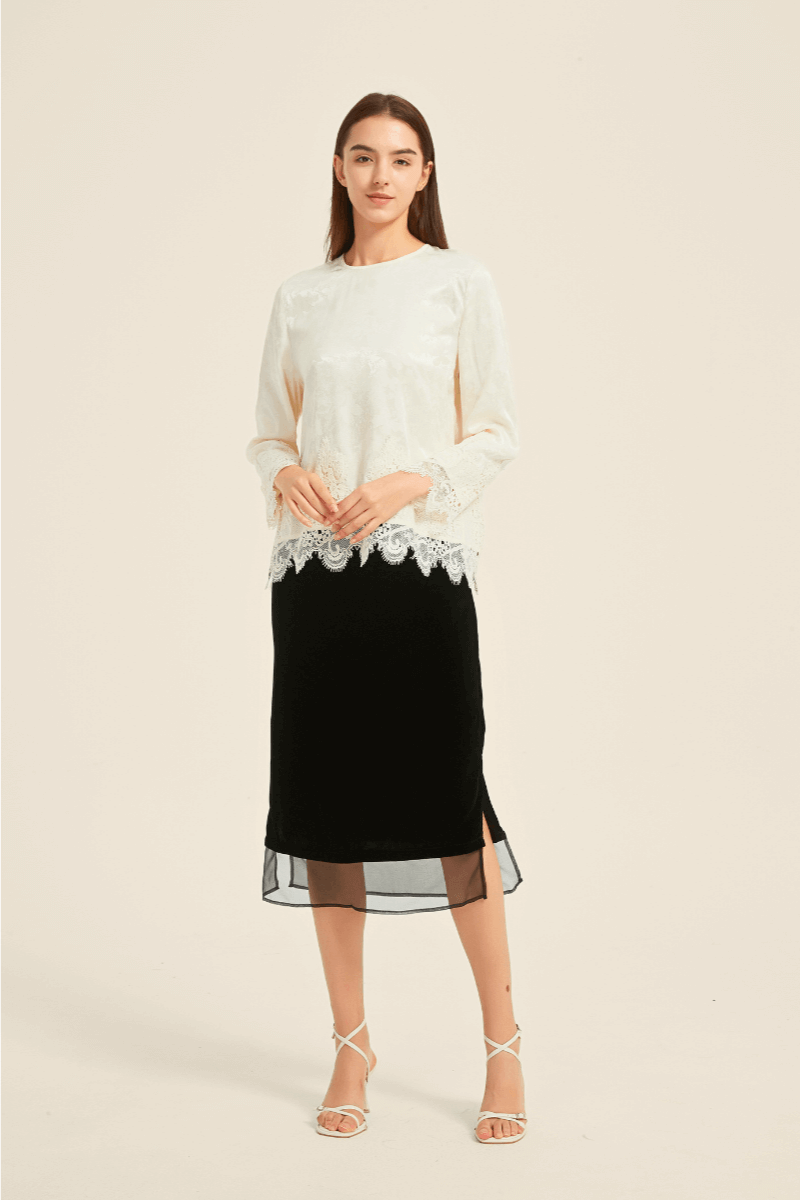 Woman wearing an ivory lace-trim long-sleeve blouse paired with a black midi skirt with side slit, styled with white strappy heels for an elegant look.