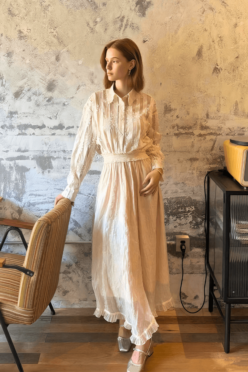 Woman wearing an ivory maxi dress with long sleeves, pleated bodice, lace waistband, and ruffle hem, styled with silver heels in a rustic indoor setting.