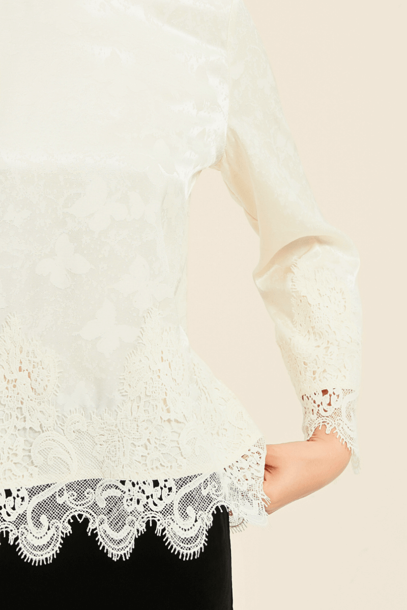 Close-up of an ivory satin blouse featuring delicate butterfly jacquard, scalloped lace trim on the hem and cuffs, paired with a black skirt.