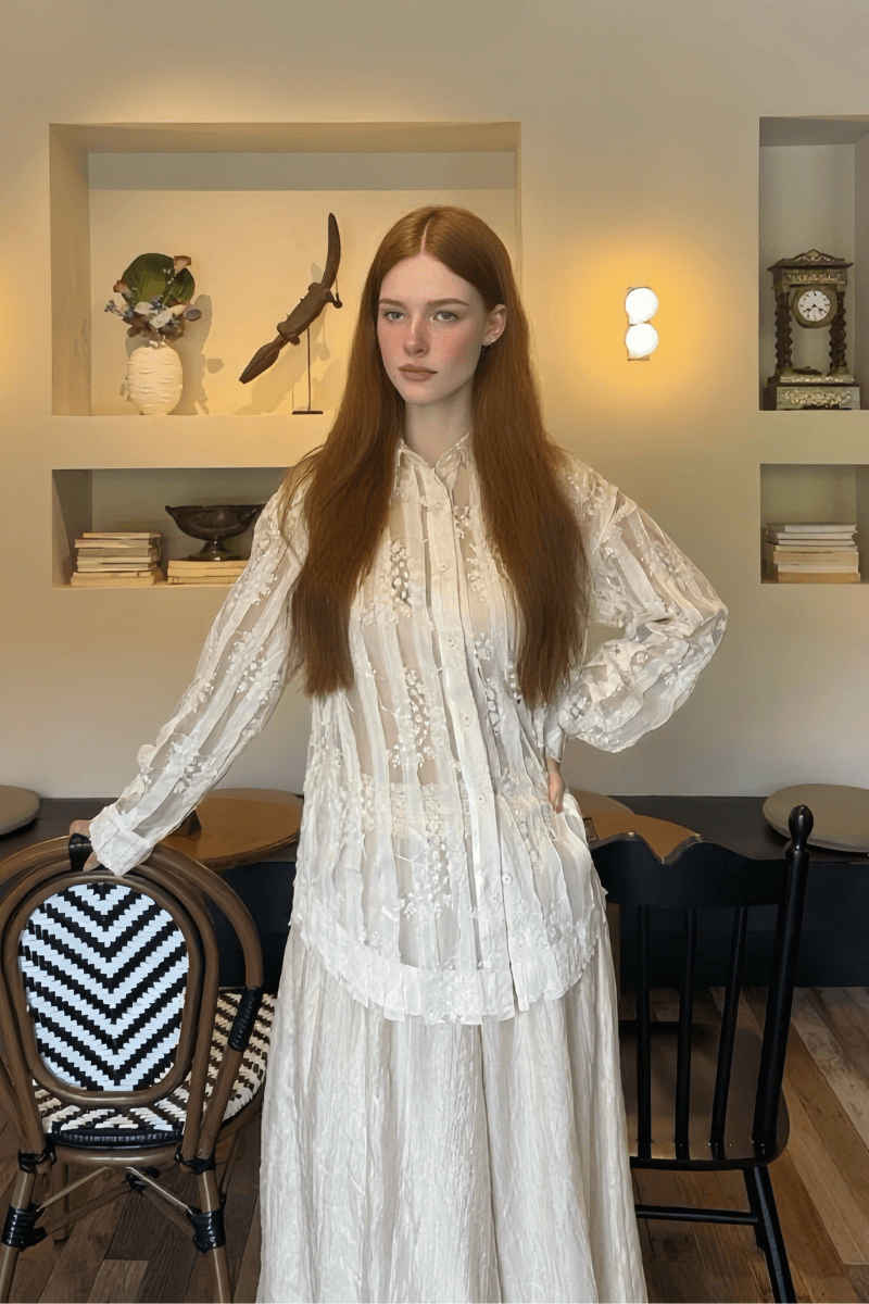 Woman wearing a sheer ivory blouse with floral embroidery, long sleeves, pleated front, and button-down closure, styled with a flowing ivory skirt in a modern indoor setting.