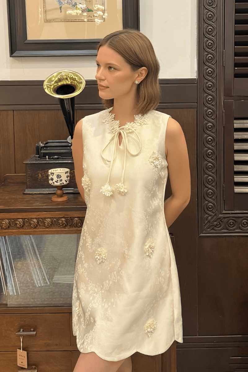 Elegant sleeveless ivory mini dress featuring a bow-tie neckline, subtle floral appliqué embellishments, and scalloped hemline, styled in a vintage-inspired interior.