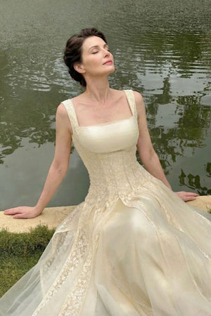 A woman in an ivory square-neck corset ballgown sits peacefully beside a lakeside edge, eyes closed, showing the gown’s textured panels and dreamy fairytale elegance.