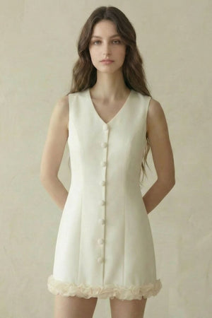 model wearing an ivory sleeveless button-front mini dress with soft rosette trim at the hem, standing against a neutral beige backdrop.