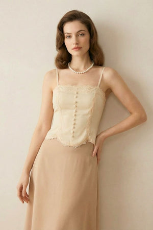 Model wearing an ivory lace-trim corset top with pearl buttons paired with a beige midi skirt, styled with soft waves and a pearl necklace in a minimal studio setting.