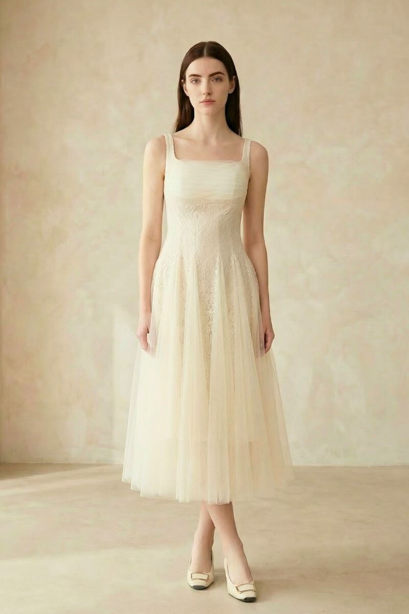 A woman wearing an ivory lace tulle midi dress with wide straps and a soft A-line silhouette, shown standing against a neutral backdrop.