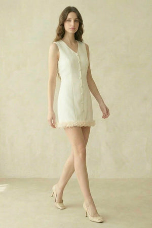 model walking in an ivory sleeveless mini dress with front buttons and rosette hem, photographed in a minimal beige studio.