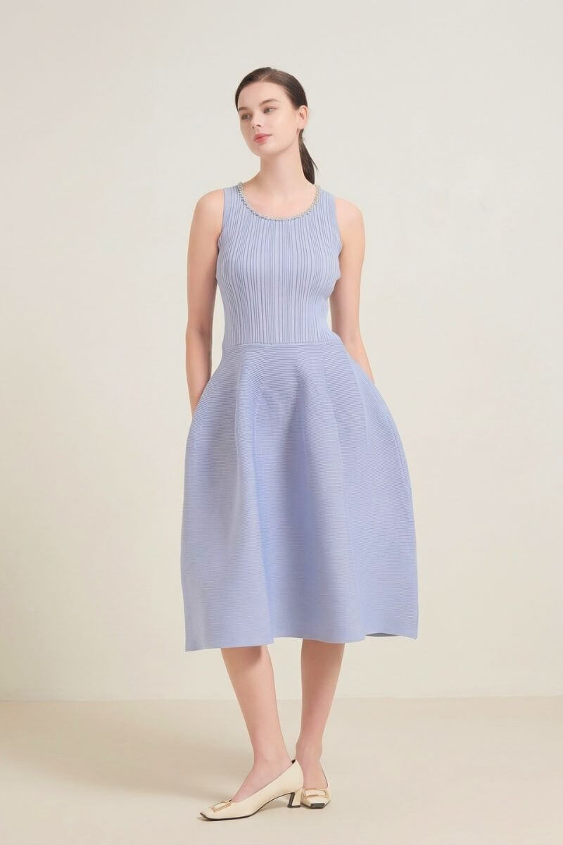 A model posing in a light blue sleeveless midi dress with a ribbed top, pearl neckline, and flared skirt, styled with cream heels in a simple studio scene.