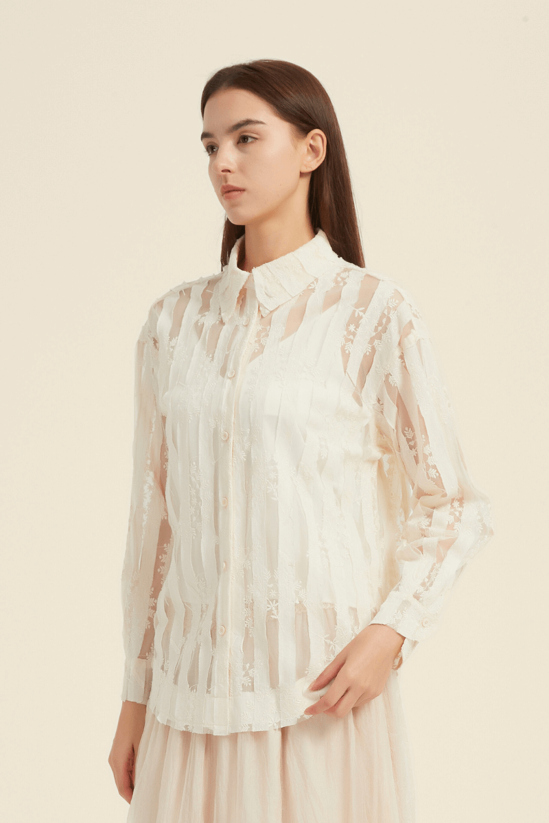 Front-facing view of a cream long-sleeve blouse with sheer striped lace panels, embroidered floral details, and a pointed collar.