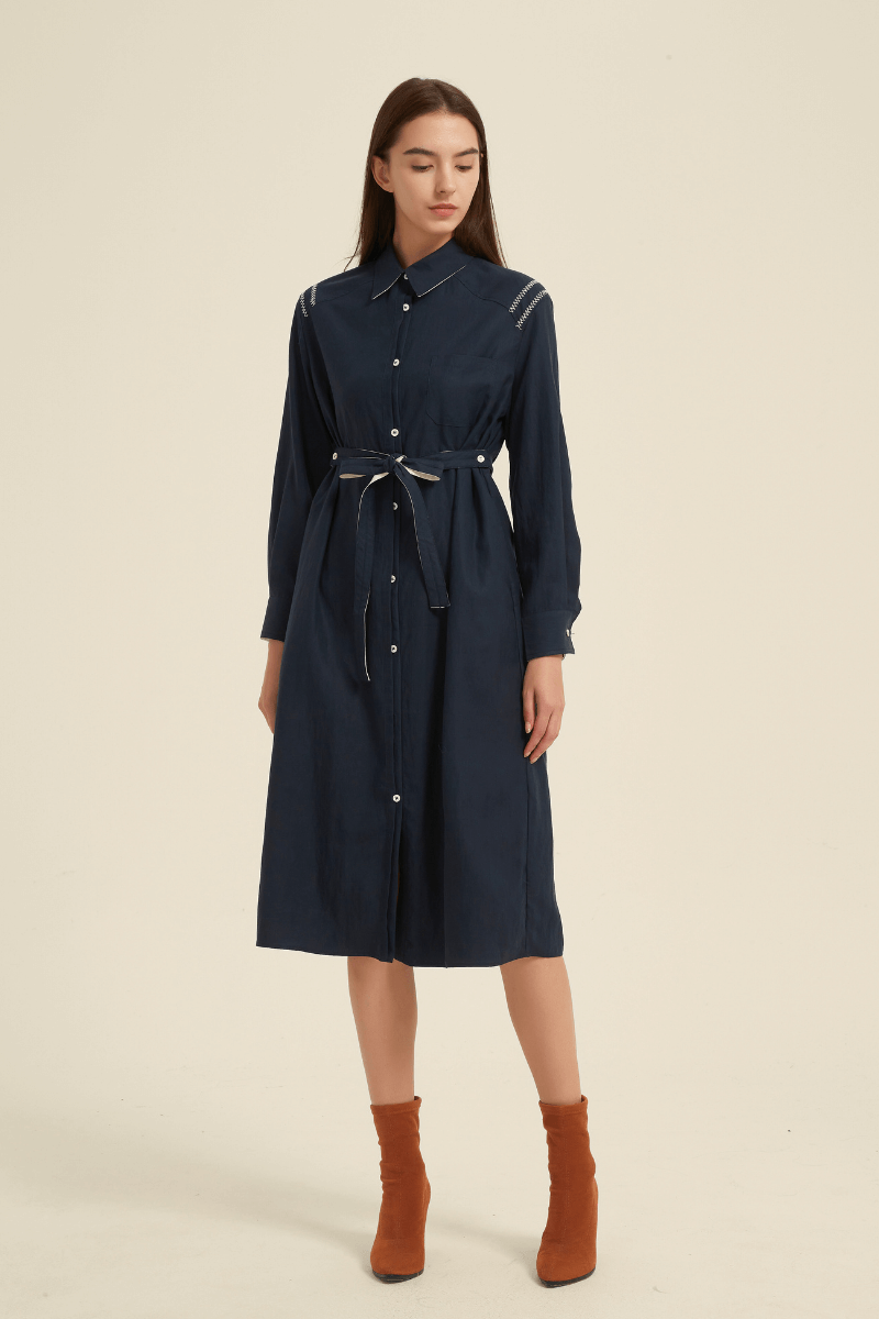 Woman wearing a navy button-down midi shirt dress with long sleeves, collared neckline, belted waist tie, and classic tailored fit.