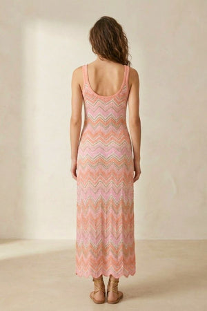 Back view of a woman wearing a pink chevron knit maxi dress with a scoop back, standing indoors in soft neutral lighting.