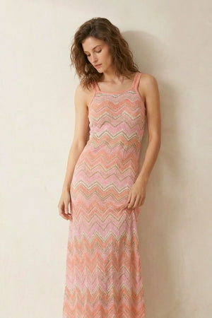 A closer front view of a woman in a pink chevron-pattern knit maxi dress, posing in a softly lit neutral studio.