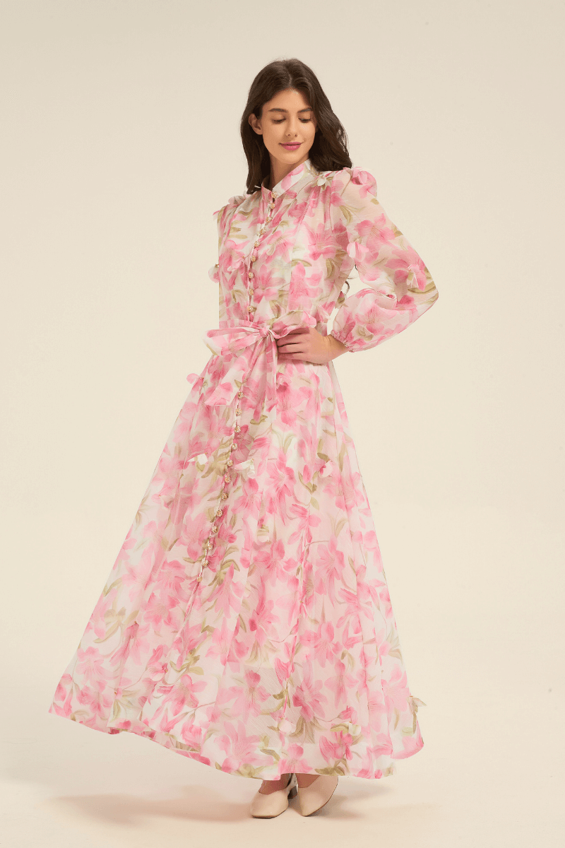Model wearing a pink floral print maxi dress with long sleeves, button-down front, and waist tie belt, styled with light heels.
