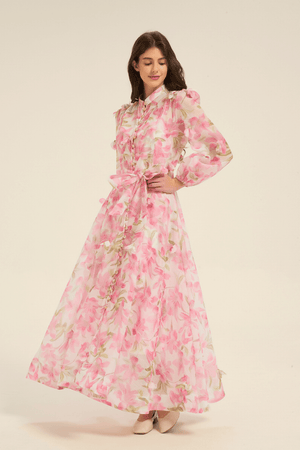 Model wearing a pink floral print maxi dress with long sleeves, button-down front, and waist tie belt, styled with light heels.