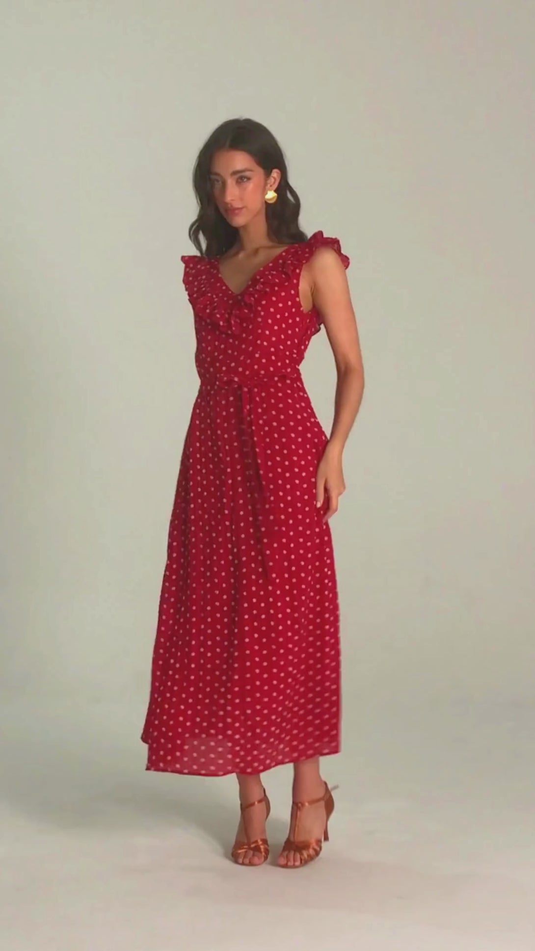 Smiling woman wearing a red ruffle polka dot midi dress with tie waist, holding the skirt hem in a light, joyful pose against a neutral background