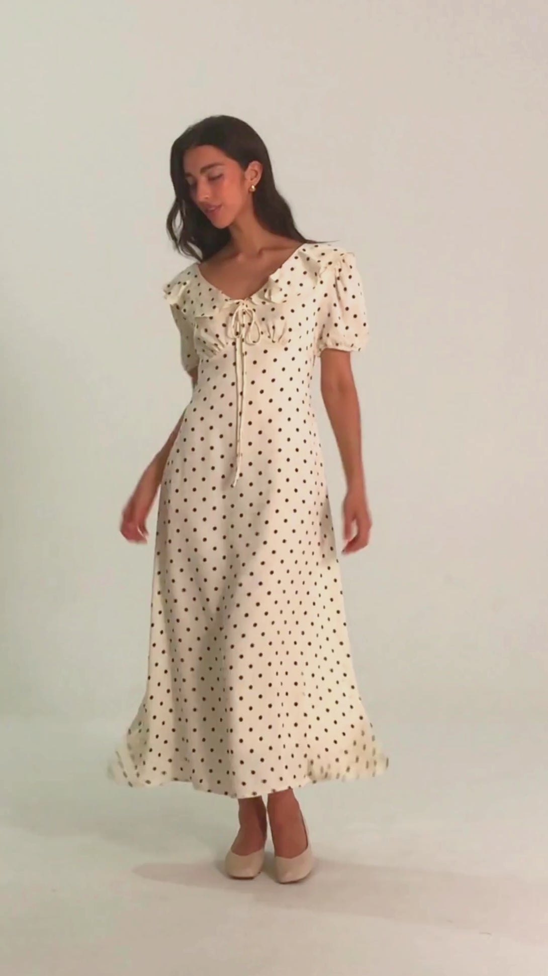 Woman wearing a white midi dress with black polka dots, puff sleeves, and a ruffled neckline with bow tie, facing forward with a slight smile